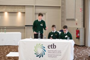 Image of three students at a table, one standing up to speak.